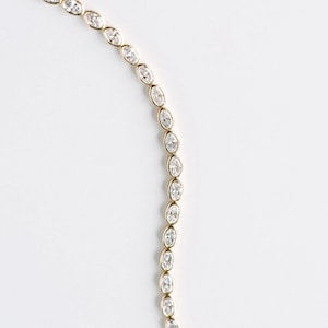 Puede incluir: Una delicada pulsera de oro con una serie de piedras preciosas claras de talla ovalada. Las piedras están engastadas muy juntas, creando una línea continua de brillo. La pulsera se muestra sobre un fondo blanco liso.