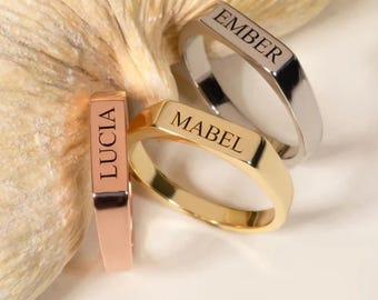 May include: Three personalized signet rings in rose gold, gold, and silver. The rings are engraved with the names "Lucia", "Mabel", and "Ember". The rings have a rectangular face and a smooth, polished finish.