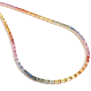 Puede incluir: Un delicado collar con un degradado de gemas coloridas. Las gemas transitan del rojo al naranja, amarillo, verde, azul y morado. El collar está engastado en un metal dorado, creando una joya vibrante y llamativa.