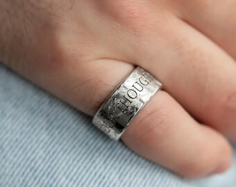 Puede incluir: Un anillo de plata con las palabras "OUR THOUGHTS" grabadas en la banda. El anillo tiene un acabado texturizado y martillado y se lleva en un dedo. El anillo es una joya.