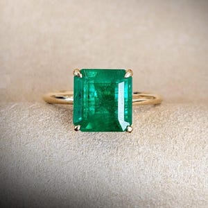 Puede incluir: Un anillo de oro con una gran esmeralda rectangular. La esmeralda es de un color verde vibrante y está engastada con cuatro garras. La banda del anillo es de oro pulido simple.