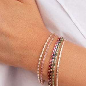 May include: Several gold bracelets, each set with a row of rectangular gemstones in various colours. The bracelets display pink, red, blue, green, and clear stones. The bracelets are worn on a wrist against a white background.