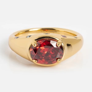 May include: A gold-colored ring with a large, oval-shaped, red gemstone as the centerpiece. The ring has a thick band and small accent stones. The gemstone is held in place by four prongs.