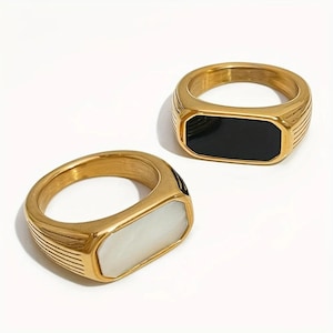 May include: Two gold signet rings. One has a black rectangular stone, the other a white rectangular stone. The rings have a ridged design on the sides and a polished finish.