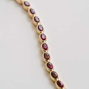 Puede incluir: Un brazalete dorado con una serie de piedras preciosas ovaladas de color rojo intenso. Cada piedra está engastada en un bisel dorado, creando una línea continua. El brazalete se muestra sobre un fondo blanco. Las piedras miden unos 0,8 cm de largo.