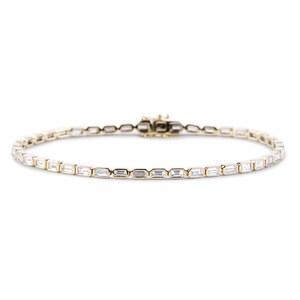Op de afbeelding: Een gouden armband met rechthoekige, heldere stenen. De armband heeft een sluiting en wordt gepresenteerd tegen een witte achtergrond. De stenen zijn gelijkmatig verdeeld en reflecteren licht.