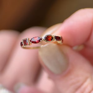 May include: A gold ring featuring a series of red gemstones in various shapes: square, oval, and round. The ring is held in a person's hand, showcasing the gemstones' vibrant color and the ring's delicate design. The ring is a piece of jewelry.