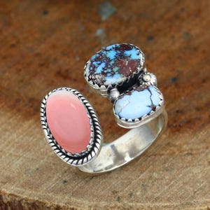 May include: A silver open-style ring featuring three oval-shaped stones. One side displays a smooth, solid pink stone, while the other side features two smaller stones: one with a dark blue and brown matrix pattern and one with a light blue colour.