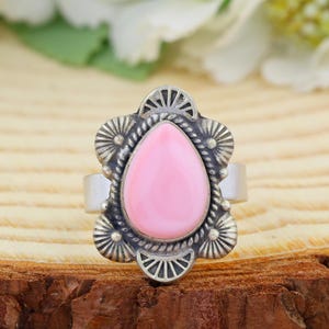 Puede incluir: Un anillo de plata con una piedra rosa lisa en forma de lágrima en el centro. El engaste de metal está decorado con un borde festoneado y patrones de líneas radiales, rodeando la piedra con una textura de cuerda trenzada y una banda pulida.