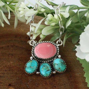 May include: A silver necklace featuring a pendant with one large oval pink stone at the top and three smaller oval turquoise stones with brown matrix patterns arranged below it, all set in twisted silver metal frames on a silver chain.