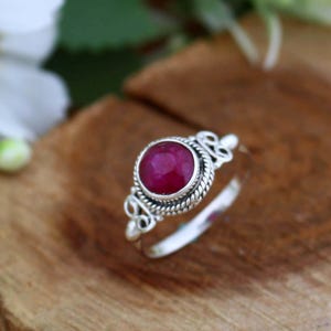 May include: A sterling silver ring featuring a large, round, deep red gemstone. The gemstone is set in a bezel setting and surrounded by a twisted rope design. The ring has decorative flourishes on the shoulders.