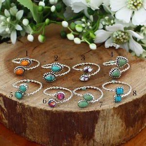 May include: A collection of silver-toned rings with various colorful stone settings, including orange, turquoise, purple, and green. The rings are displayed on a rustic wooden surface, surrounded by white flowers and greenery.