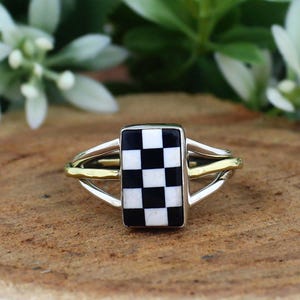 May include: A silver ring with a rectangular black and white checkered design. The ring has a gold band that connects to the silver band. The ring is set on a wooden surface with green foliage in the background.