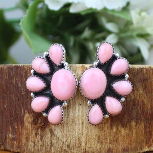 Könnte beinhalten: Ein Paar rosa Edelstein-Ohrstecker. Jeder Ohrring hat einen zentralen ovalen Stein, der von kleineren, tropfenförmigen Steinen umgeben ist. Die Steine sind in einem silberfarbenen Metall mit dunkler, oxidierter Oberfläche gefasst.