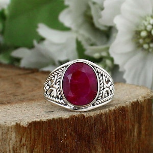 Puede incluir: Anillo de plata con una gema ovalada de color rojo intenso. El anillo tiene un diseño ornamentado y texturizado alrededor de la gema. El anillo está colocado sobre un trozo de madera, con flores blancas al fondo.