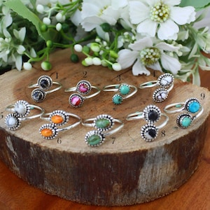 May include: A collection of nine silver rings, each featuring two oval-shaped stones in various colors, including black, white, turquoise, orange, and green. The rings are displayed on a wooden surface, with white flowers in the background.