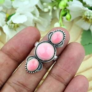 Op de afbeelding: Een zilveren ring met drie gladde, ovaalvormige roze stenen in een verticale rij. De middelste steen is de grootste, geflankeerd door twee kleinere stenen, allemaal gezet in decoratief zilverwerk met gedetailleerde lijnen.