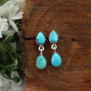 Puede incluir: Un par de pendientes colgantes turquesa con engaste plateado. Cada pendiente presenta dos piedras turquesas en forma de lágrima conectadas por una pequeña cadena. Los pendientes se exhiben sobre una superficie de madera.