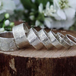 Puede incluir: Una colección de anillos de plata con patrones geométricos, que varían en ancho de 2 mm a 14 mm. Los anillos se muestran sobre una superficie de madera, con un fondo floral borroso.
