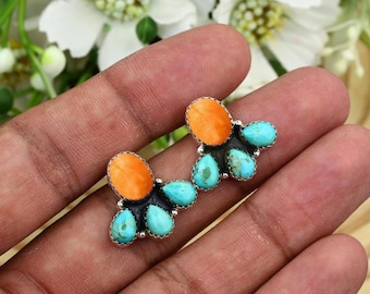 May include: A cluster-style ring featuring two prominent oval orange stones and six smaller teardrop-shaped turquoise stones set in a dark metal frame. The stones are arranged in a balanced, organic pattern with serrated metal bezels.