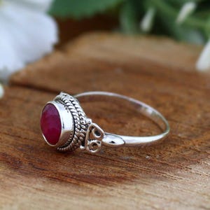 May include: A silver ring featuring a round, deep red gemstone. The ring has an ornate, textured setting around the stone and a simple, polished band. The ring is displayed on a wooden surface.