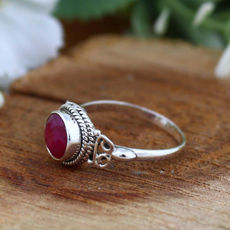 May include: A silver ring featuring a round, deep red gemstone. The ring has an ornate, textured setting around the stone and a simple, polished band. The ring is displayed on a wooden surface.