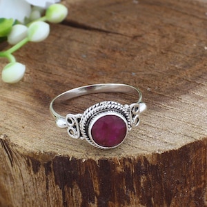 Op de afbeelding: Een zilveren ring met een grote, ronde, dieprode edelsteen in het midden. De edelsteen is gezet in een decoratieve zilveren zetting met een gevlochten textuur. De ring heeft een dunne band met sierlijke details aan weerszijden van de edelsteen.