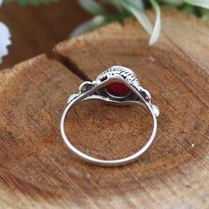 May include: A silver ring with a round, deep red gemstone. The ring has an ornate, textured setting and a delicate band. The gemstone is the focal point, set against a backdrop of natural wood grain.
