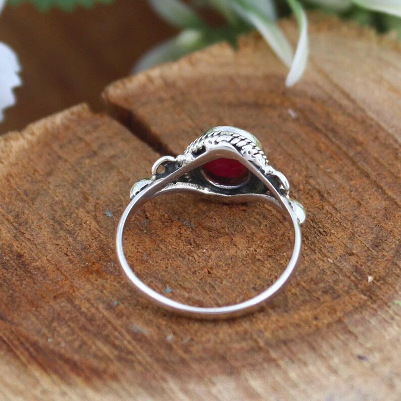 May include: A silver ring with a round, deep red gemstone. The ring has an ornate, textured setting and a delicate band. The gemstone is the focal point, set against a backdrop of natural wood grain.