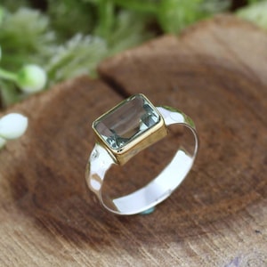 May include: A silver ring featuring a rectangular, pale green gemstone set within a gold bezel. The ring's band has a hammered texture, and the gemstone is the focal point. The ring is displayed on a wooden surface.