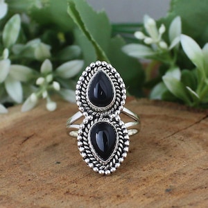 May include: A silver ring with two black, teardrop-shaped gemstones. The stones are set within a beaded frame. The ring is shown on a wooden surface, with green leaves and white flowers in the background.