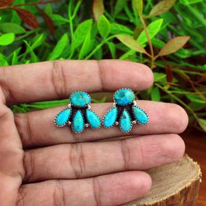 Puede incluir: Un par de pendientes de botón de color turquesa. Cada pendiente presenta un grupo de piedras turquesas en un diseño floral, engastadas en metal plateado. Los pendientes se muestran en una mano.