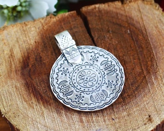 May include: A silver pendant with a circular design featuring intricate patterns and a decorative bail. The pendant has a detailed floral center and a scalloped edge. The bail has a geometric design.