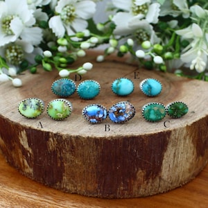 May include: A collection of gemstone stud earrings displayed on a wooden surface. The earrings feature various colors, including turquoise, green, and blue, with silver-colored metal settings. The earrings are arranged in pairs, showcasing their unique designs and textures.