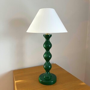 May include: A green table lamp with a white lampshade. The lamp has a glossy finish and a stacked, rounded base. The lampshade is cone-shaped. The lamp is on a wooden surface.
