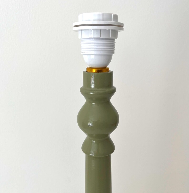 May include: A green lamp base with a white lamp holder. The base has a decorative, turned design and a gold-colored accent. The lamp holder is white and has a ribbed texture. The lamp base is painted in a muted green color.
