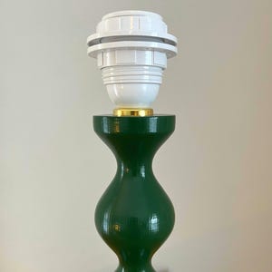 May include: A green table lamp base with a white lamp holder. The base has a curvy, hourglass shape. The lamp holder is white with a gold-coloured accent at the base.