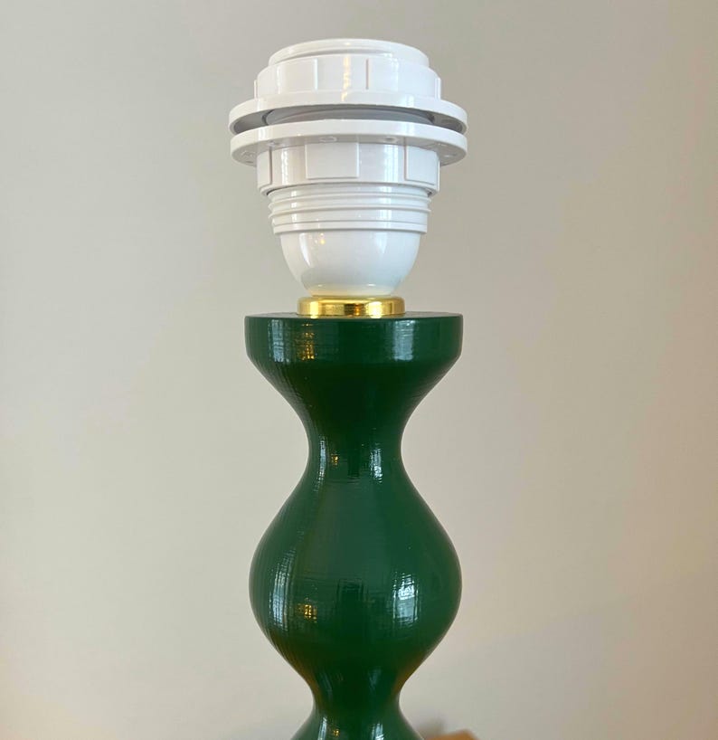 May include: A green table lamp base with a white lamp holder. The base has a curvy, hourglass shape. The lamp holder is white with a gold-coloured accent at the base.