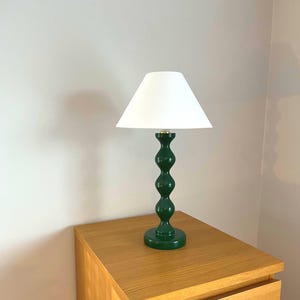 May include: A green table lamp with a white lampshade. The lamp has a decorative, rounded base and a dark green, stacked-ball design on the stem. The lamp is sitting on a wooden bedside table.