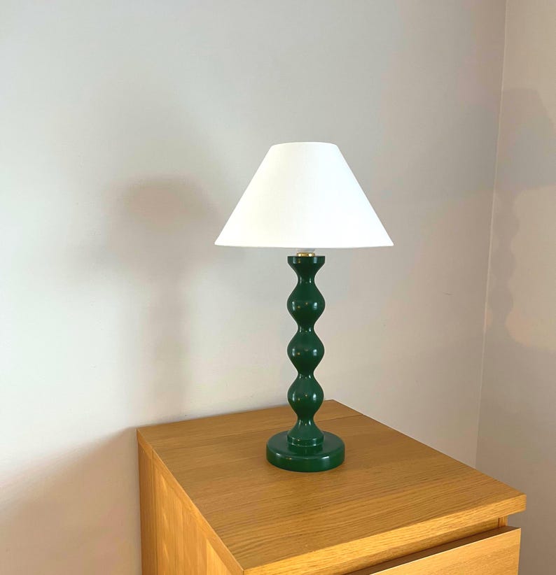 May include: A green table lamp with a white lampshade. The lamp has a decorative, rounded base and a dark green, stacked-ball design on the stem. The lamp is sitting on a wooden bedside table.