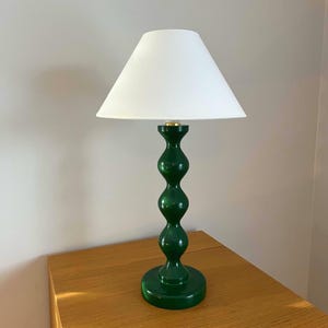 May include: A green table lamp with a white lampshade. The lamp has a glossy finish and a unique, stacked-orb design. The base is round, and the shade is a classic cone shape. The lamp is on a wooden surface.