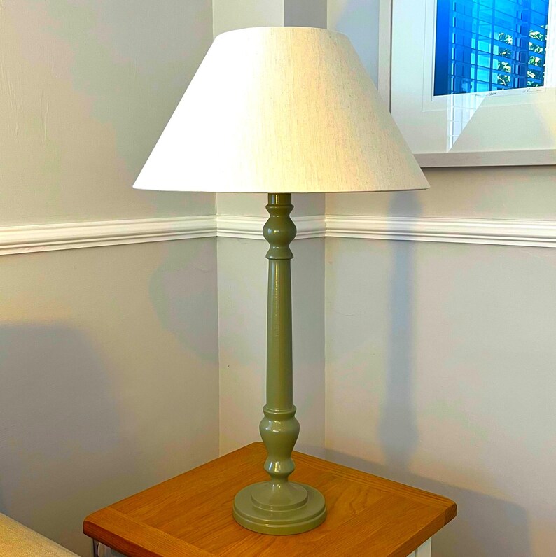 May include: A green table lamp with a beige lampshade. The lamp has a decorative, turned base and a long, slender stem. The lampshade is conical. The lamp is sitting on a wooden table.
