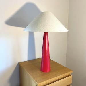 May include: A contemporary table lamp featuring a vibrant red conical base and a light grey lampshade. The lamp is placed on a light wood bedside table. The lamp's shadow is cast on the wall. This is a decorative lighting item.