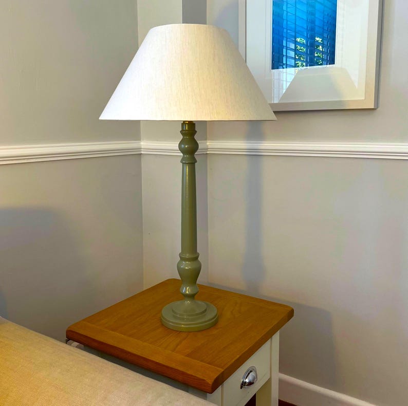 May include: A tall, olive green table lamp with a cream-colored lampshade. The lamp sits on a small, square wooden side table with a drawer. The lamp's base has a decorative, turned design.