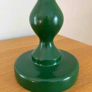 May include: A dark green wooden candlestick holder with a glossy finish. The base is a wide, flat circle, supporting a shapely, bulbous body that tapers to a narrower top. The surface reflects light, showing the wood grain.
