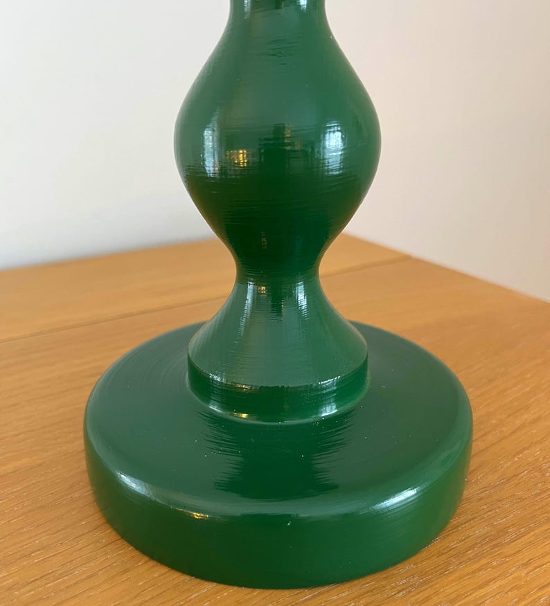 May include: A dark green wooden candlestick holder with a glossy finish. The base is a wide, flat circle, supporting a shapely, bulbous body that tapers to a narrower top. The surface reflects light, showing the wood grain.