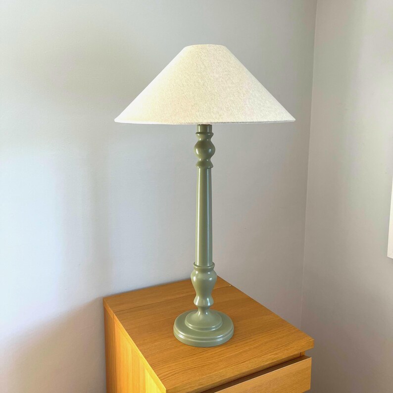 May include: A sage green table lamp with a cream-colored lampshade. The lamp has a decorative, turned base and a long, slender stem. The lampshade is conical. The lamp is sitting on a wooden nightstand.