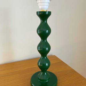 May include: A green table lamp with a white bulb socket. The lamp has a rounded, hourglass-shaped body and a circular base. The lamp is made of wood and has a glossy finish. The lamp is approximately 38 cm tall.