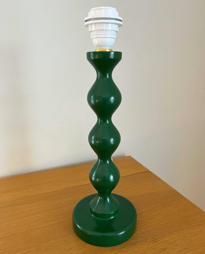 May include: A green table lamp with a white bulb socket. The lamp has a rounded, hourglass-shaped body and a circular base. The lamp is made of wood and has a glossy finish. The lamp is approximately 38 cm tall.