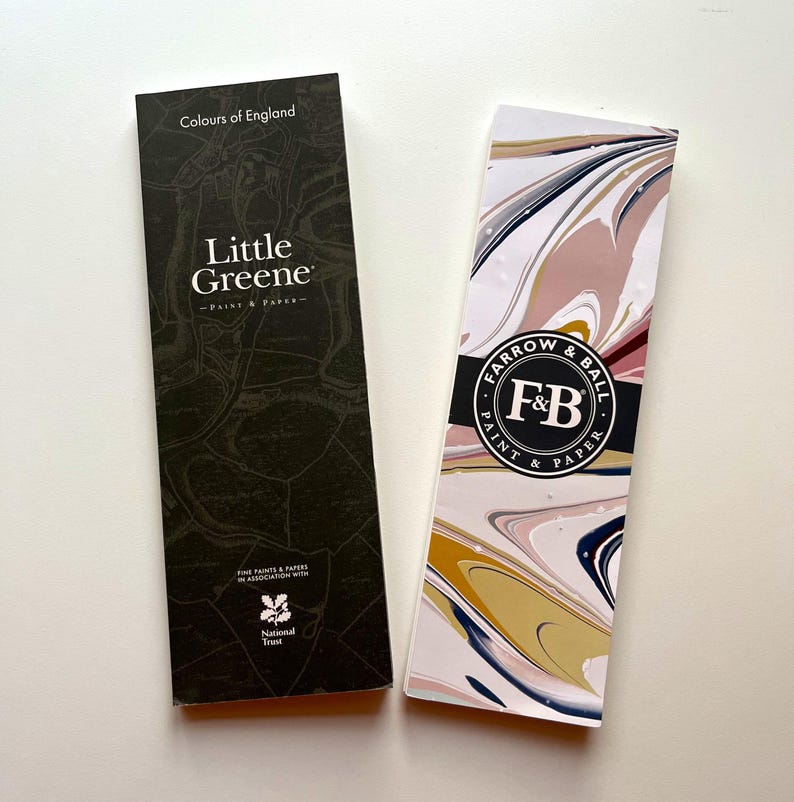 May include: Two paint and paper sample cards. One is dark with the text "Colours of England" and "Little Greene - Paint & Paper". The other has a marbled design with the text "Farrow & Ball Paint & Paper".